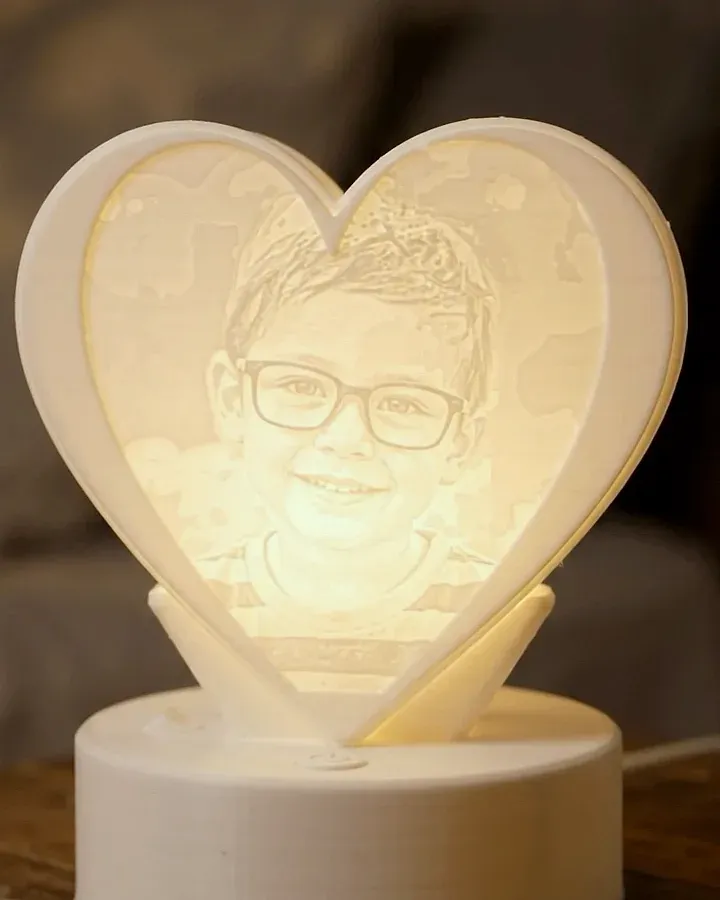 Une lithophanie en forme de cœur allumée sur une table de chevet, la photo d’un enfant à lunettes apparaît dans le plastique blanc, posée sur un socle lumineux rond